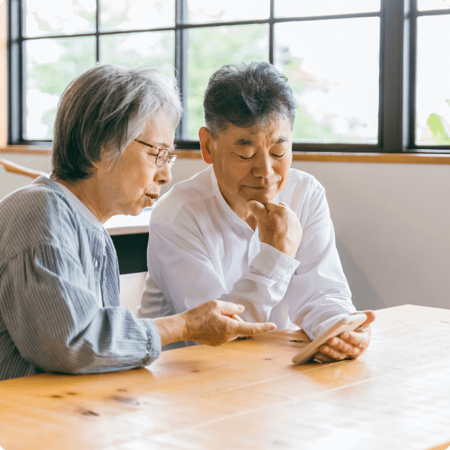 中年の夫婦がスマートフォンを見ながら話し合っている場面。背景には窓がある。